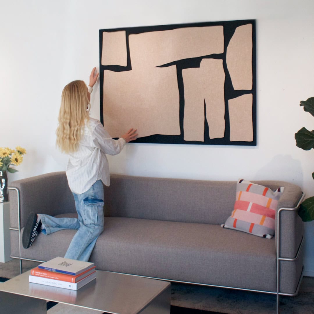 Woman mounting abstract decorative sound panel above grey sofa in modern living room with soft lighting